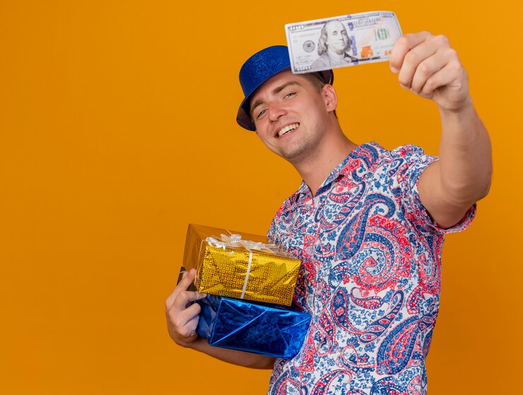 Foto grátis garoto jovem sorridente com chapéu azul, segurando caixas de presente e segurando dinheiro para a câmera, isolado em um fundo laranja com espaço de cópia