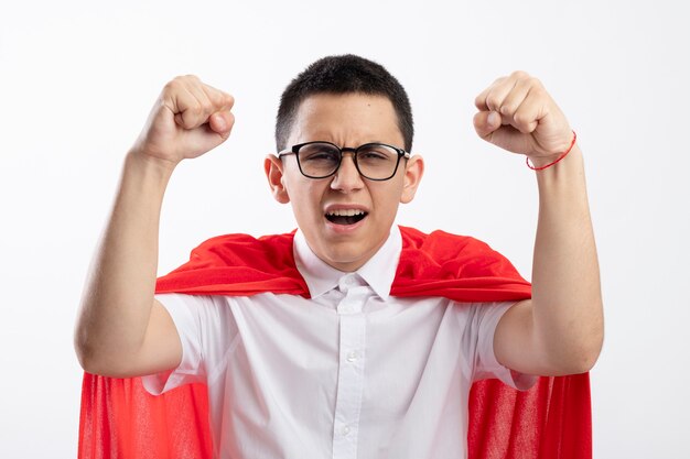 Garoto jovem super-herói alegre com capa vermelha usando óculos, olhando para a câmera, levantando os punhos e aproveitando a vitória isolada no fundo branco