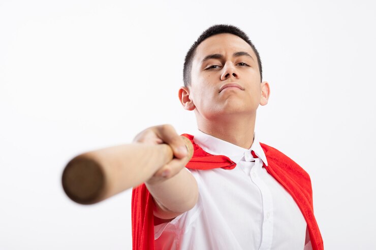 Foto grátis garoto jovem super-herói confiante com capa vermelha esticando o taco de beisebol em direção à câmera, olhando para a câmera isolada no fundo branco