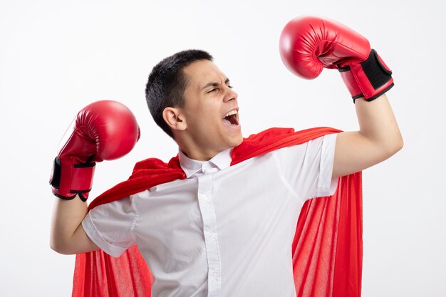 Garoto jovem super-herói confiante com capa vermelha usando luvas de boxe fazendo um gesto forte olhando para a mão dele gritando isolado no fundo branco