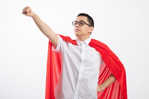 Garoto jovem super-herói confiante com capa vermelha usando óculos, segurando a mão na cintura, olhando para o lado esticando o punho isolado no fundo branco