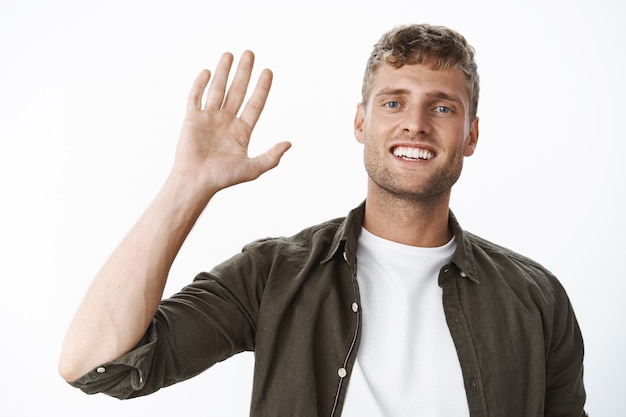 Garoto loiro amigável dizendo olá acenando amigavelmente na frente de saudação, sorrindo amplamente, estando de bom humor, posando calmo e despreocupado contra a parede cinza como dizendo oi para os amigos