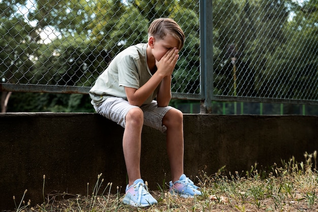Foto grátis garoto triste de tiro completo sentado ao ar livre