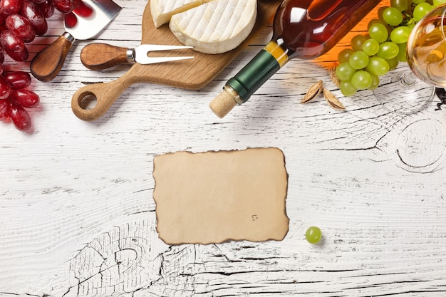 Garrafa de vinho branca, uva, mel, queijo, copo de vinho e folha de papel na placa de madeira branca. vista superior com espaço de cópia.