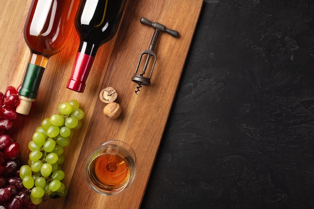 Garrafas de vinho tinto e branco com cacho de uvas, nozes, saca-rolhas e um copo de vinho na placa de madeira e fundo preto. vista superior com espaço de cópia.