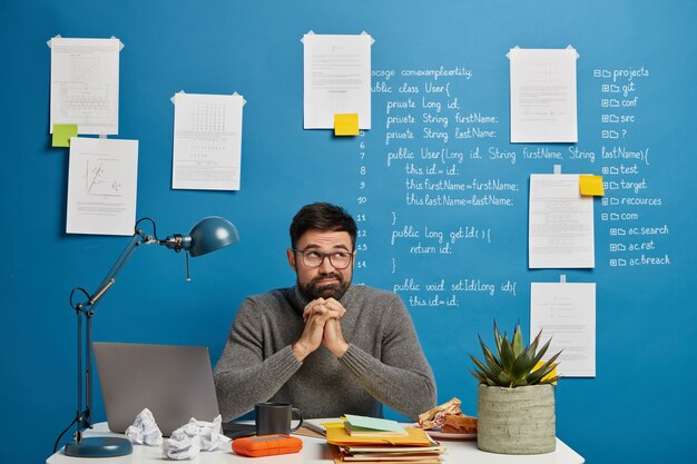 Geek masculino profissional sério concentrado no monitor de um laptop moderno, usa óculos ópticos, posa no espaço de coworking contra um fundo azul