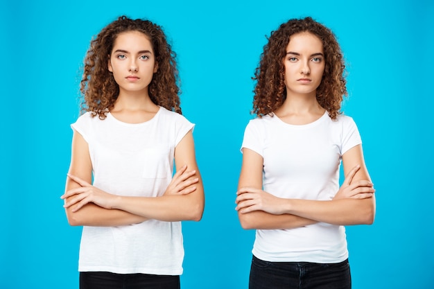 Gêmeos de duas meninas posando com os braços cruzados sobre parede azul