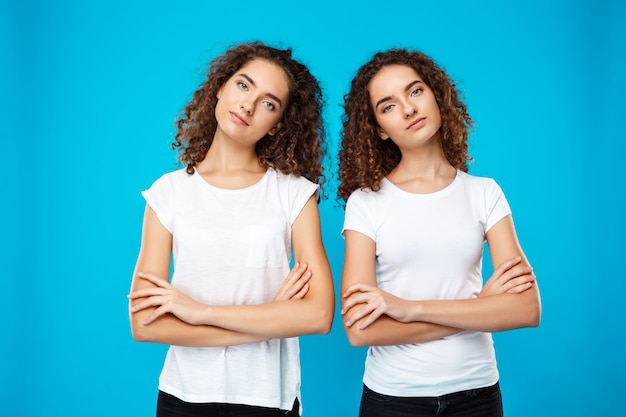 Gêmeos de duas meninas posando com os braços cruzados sobre parede azul