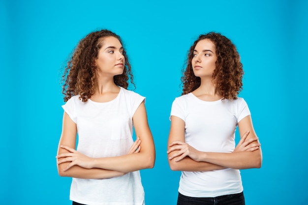 Gêmeos de duas meninas posando com os braços cruzados sobre parede azul