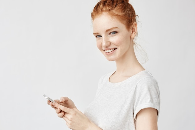 Gengibre mulher sorrindo segurando telefone twitting ou usando mídias sociais na parede branca