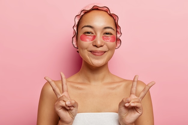 Gentil senhora oriental gesticula com as duas mãos, mostra o símbolo da paz, cuida da pele com tapa-olhos, usa touca de banho para evitar que o cabelo fique molhado, enrolada em toalha branca. cosmetologia