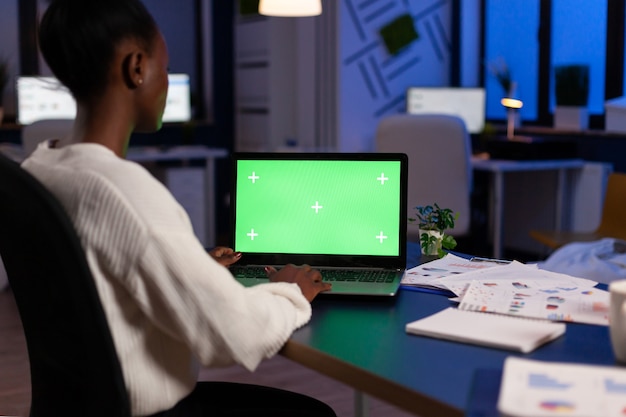 Gerente africano digitando em um laptop com visor de chroma key durante a noite no escritório de inicialização trabalhando horas extras