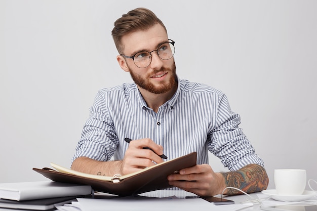 Gerente atencioso com óculos redondos, usa camisa formal, escreve no caderno enquanto está sentado no local de trabalho,