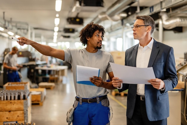 Gerente da empresa segurando planos de projeto enquanto conversa com o trabalhador manual afro-americano que está apontando para algo na fábrica