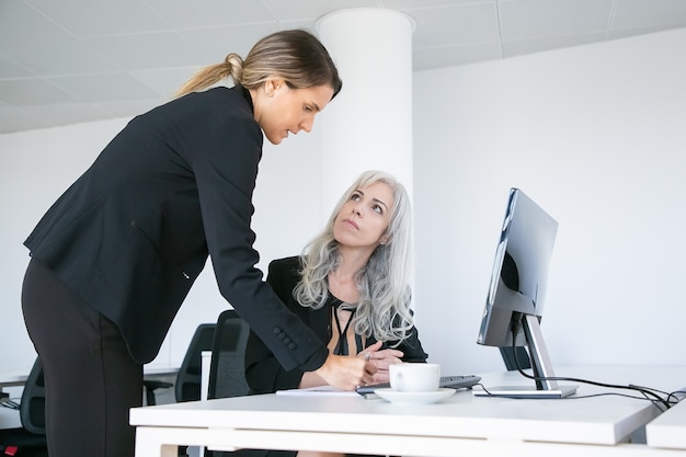 Foto grátis gerente de projeto afixando assinatura no relatório dos funcionários. colegas de trabalho femininas sentadas e em pé no local de trabalho com monitor e xícara de café. conceito de comunicação empresarial