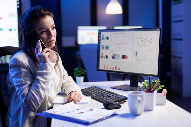 Gerente estressado falando no smartphone com o funcionário, trabalhando em problemas financeiros antes dos prazos, sentado à mesa no escritório comercial