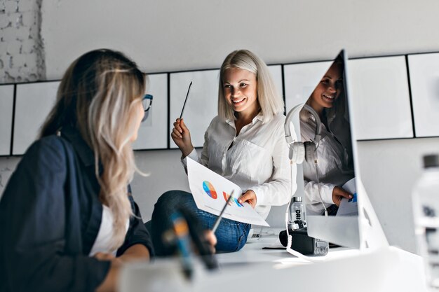 Gerente feminina loira sorridente segurando infográfico e lápis, enquanto está sentado na mesa. Retrato interno de duas mulheres trabalhando com computador no escritório.