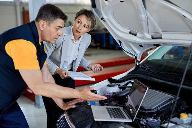 Gerente feminino e mecânico de automóveis cooperando ao usar o laptop e testando o desempenho do motor na oficina de automóveis