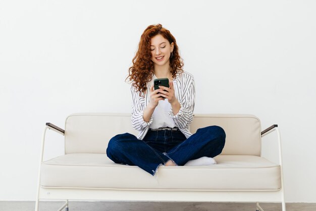 Ginger jovem deitada no sofá usando aplicativo on-line no smartphone moderno, passando o fim de semana em casa