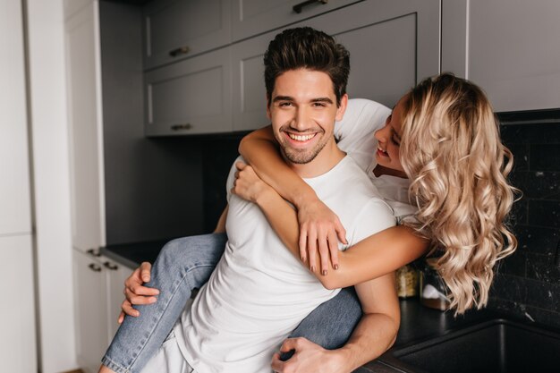 Glamourosa garota branca abraçando o marido com um sorriso. Retrato interno de casal posando na cozinha pela manhã.