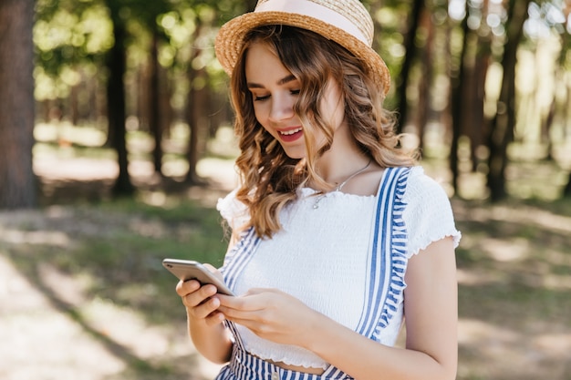 Glamourosa garota encaracolada com roupas da moda, olhando para a tela do telefone. Tiro ao ar livre da fascinante modelo feminino em mensagem de texto de chapéu após photoshoot no parque.