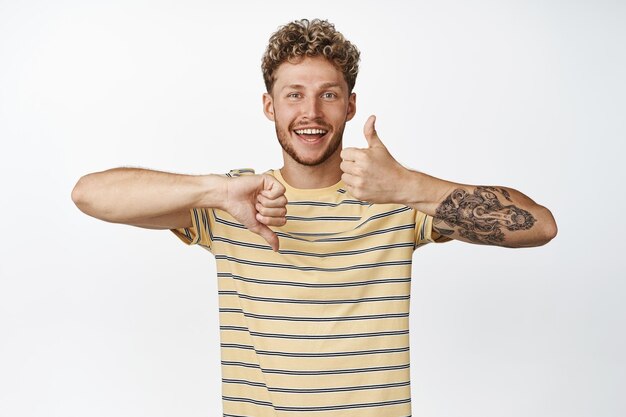 Gostar ou não gostar de um loiro feliz sorridente, mostrando os polegares para cima e os polegares para baixo, gestos prós ou contras de pé na camiseta sobre fundo branco