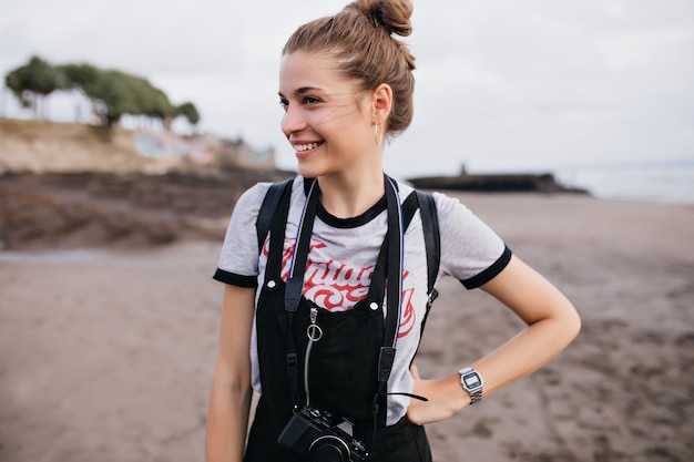 Foto grátis graciosa fotógrafa em pose confiante na praia. menina agradável num relógio de pulso moderno, sorrindo na natureza.