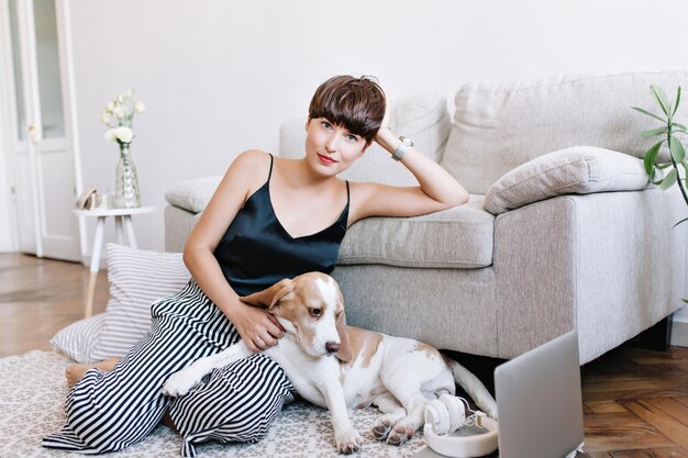 Graciosa garota de cabelos castanhos em um top preto relaxando no carpete perto de almofadas listradas e acariciando o filhote de cachorro beagle