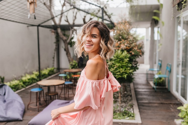 Graciosa garota de cabelos curtos olhando por cima do ombro em um aconchegante restaurante de rua. Foto ao ar livre da agradável senhora bronzeada dançando no vestido rosa.
