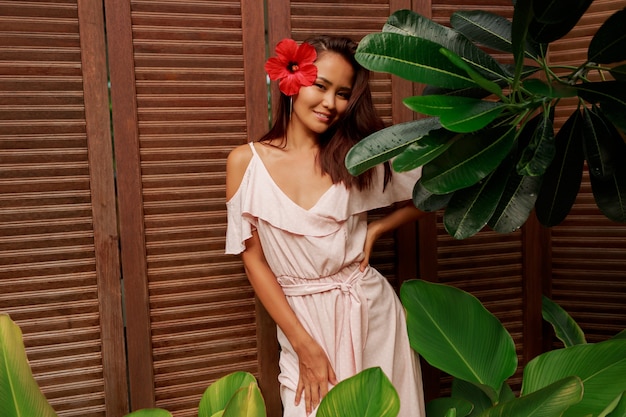 Graciosa mulher asiática com pele perfeita e flor de hibisco nos cabelos posando sobre parede de madeira