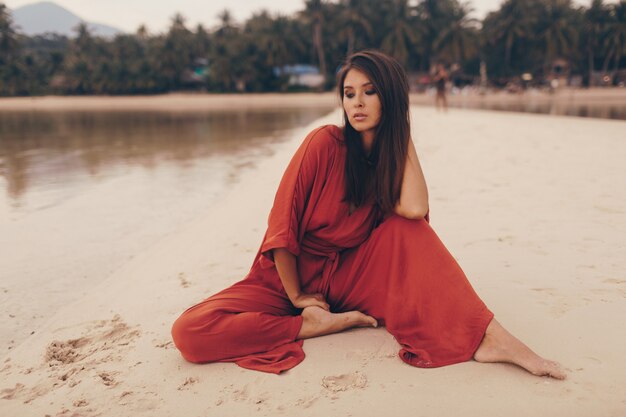 Graciosa mulher posando na praia, sentado na areia com vestido vermelho