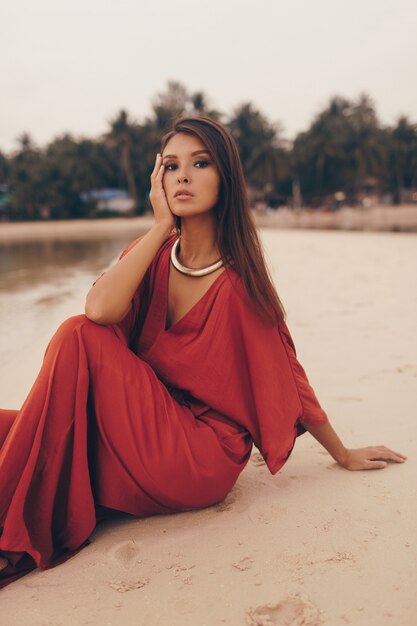 Graciosa mulher posando na praia, sentado na areia com vestido vermelho