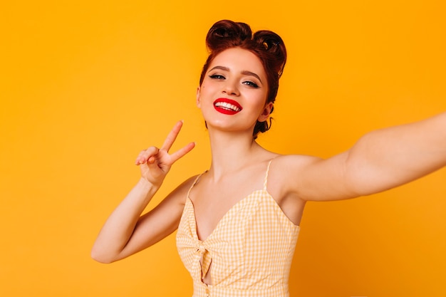 Graciosa senhora pinup mostrando o símbolo da paz. foto de estúdio de garota feliz com maquiagem brilhante, tomando selfie no espaço amarelo.