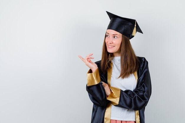 Graduada feminina apontando para o lado em uniformes, roupas casuais e olhando alegre. vista frontal.