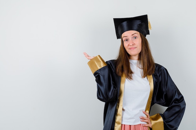 Foto grátis graduada feminina em traje acadêmico, fingindo mostrar algo e parecendo hesitante, vista frontal.