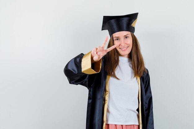 Graduada feminina mostrando gesto de vitória em roupas casuais e uniformes e olhando confiante, vista frontal.