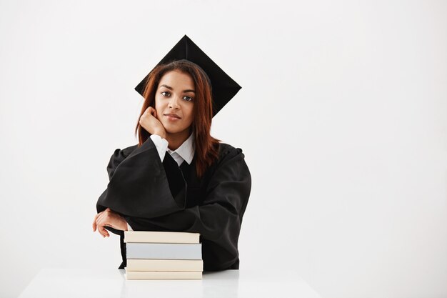 Graduado feminino Africano em cap e manto sorrindo sentado com livros.