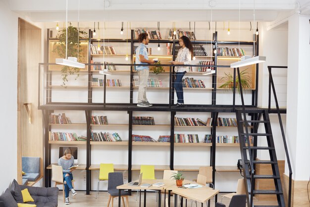 Grande biblioteca moderna com design minimalista, computadores, cadeiras confortáveis, segundo andar com estantes de livros. Manhã em local tranquilo e acolhedor. Pessoas gastando tempo na biblioteca da universidade.