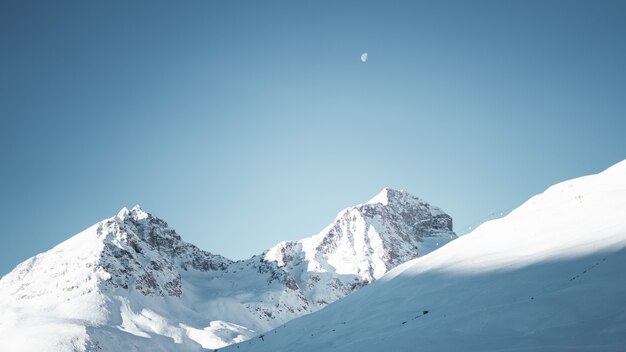 Grande plano de montanhas cobertas de neve sob um céu azul claro com meia-lua