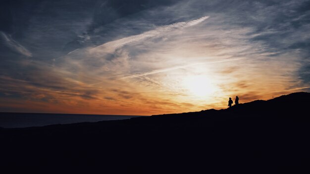 Grande plano de silhuetas de duas pessoas andando em uma colina com o sol brilhando