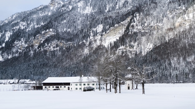 Grande plano de uma casa branca, rodeada por árvores e montanhas cobertas de neve