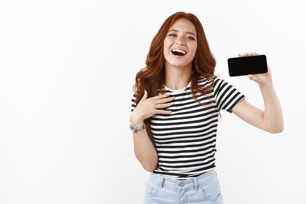 Grata e lisonjeada ruiva fofa em camiseta listrada mostrando a tela do smartphone tocando horizontalmente o peito grata e tocada sorrindo alegremente rindo e olhando para a câmera