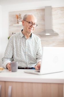 Grayhaired sênior aposentado homem navegando na internet. vida diária do homem sênior na cozinha durante o café da manhã, usando laptop segurando uma xícara de café. idoso aposentado trabalhando em casa, teletrabalho com você