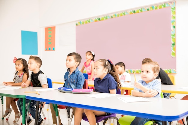 Grupo de alunos da pré-escola prestando atenção ao professor e parecendo interessados em uma sala de aula