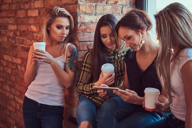 Grupo de amigas em roupas casuais discutindo enquanto procura algo em um tablet digital em uma sala com um interior loft.