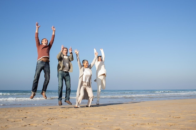 Grupo de amigos seniores animados se divertindo na praia em dia ensolarado de outono. Homens e mulheres rindo e pulando alto enquanto levantam os braços em triunfo. Férias, alegria, conceito de amigos