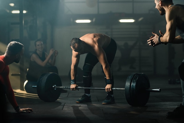 Grupo de atletas apoiando o homem de construção muscular que está realizando levantamento terra com barra em uma academia