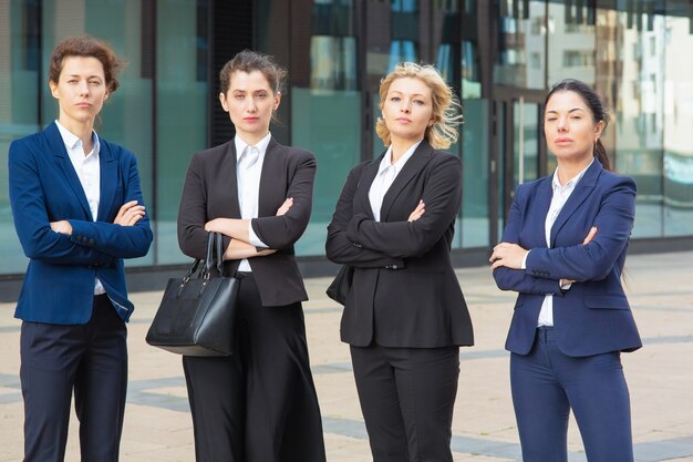 Grupo de empresárias sérias com os braços cruzados em pé juntos perto do prédio de escritórios, posando, olhando para a câmera. Vista frontal. Equipe de negócios ou conceito de trabalho em equipe