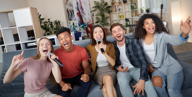 Grupo de entretenimento doméstico de jovens amigos multiculturais felizes jogando karaokê em casa e cantando com