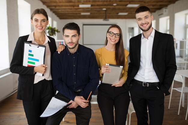 Grupo de jovens empresários em ternos clássicos olhando alegremente para a câmera juntos enquanto passam o tempo no trabalho no escritório moderno